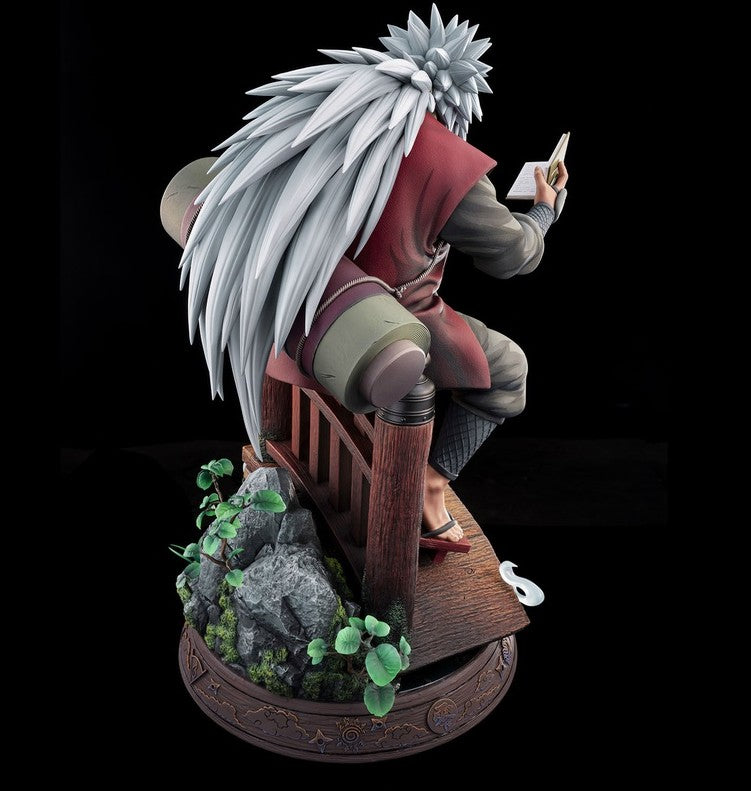 A detailed collectible statue of the anime character Madara Uchiha seated on a wooden chair, holding an open book. The figure features long white hair, a red and gray outfit, and is mounted on an ornate circular base with rocks and green plants, set against a solid black background.