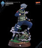 A detailed collectible figurine of Kakashi Hatake from Naruto, posed dynamically on a decorative tree stump base with swirling smoke effects. The figure is dressed in his signature blue and green ninja attire, standing on a brown carved base against a solid black background.