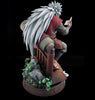 A detailed collectible statue of the anime character Madara Uchiha seated on a wooden chair, holding an open book. The figure features long white hair, a red and gray outfit, and is mounted on an ornate circular base with rocks and green plants, set against a solid black background.