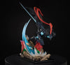 A detailed collectible anime figurine of a dark armored warrior with a glowing blue sword and flowing red hair, posed on a rocky base with blue and red energy effects, set against a black background.