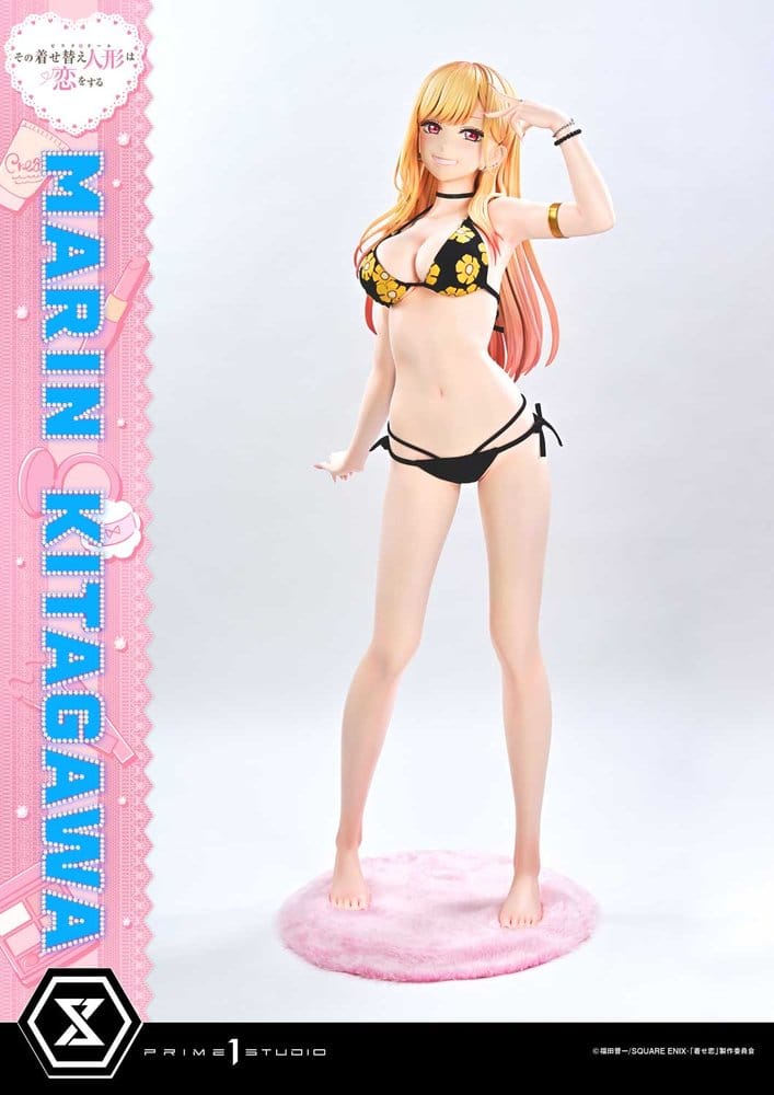 A full-body anime figurine of Marin Kitagawa wearing a black bikini with yellow floral print, standing on a pink round base against a light gray studio background. A pink vertical banner on the left reads 'MARIN KITAGAWA' with branding at the bottom.